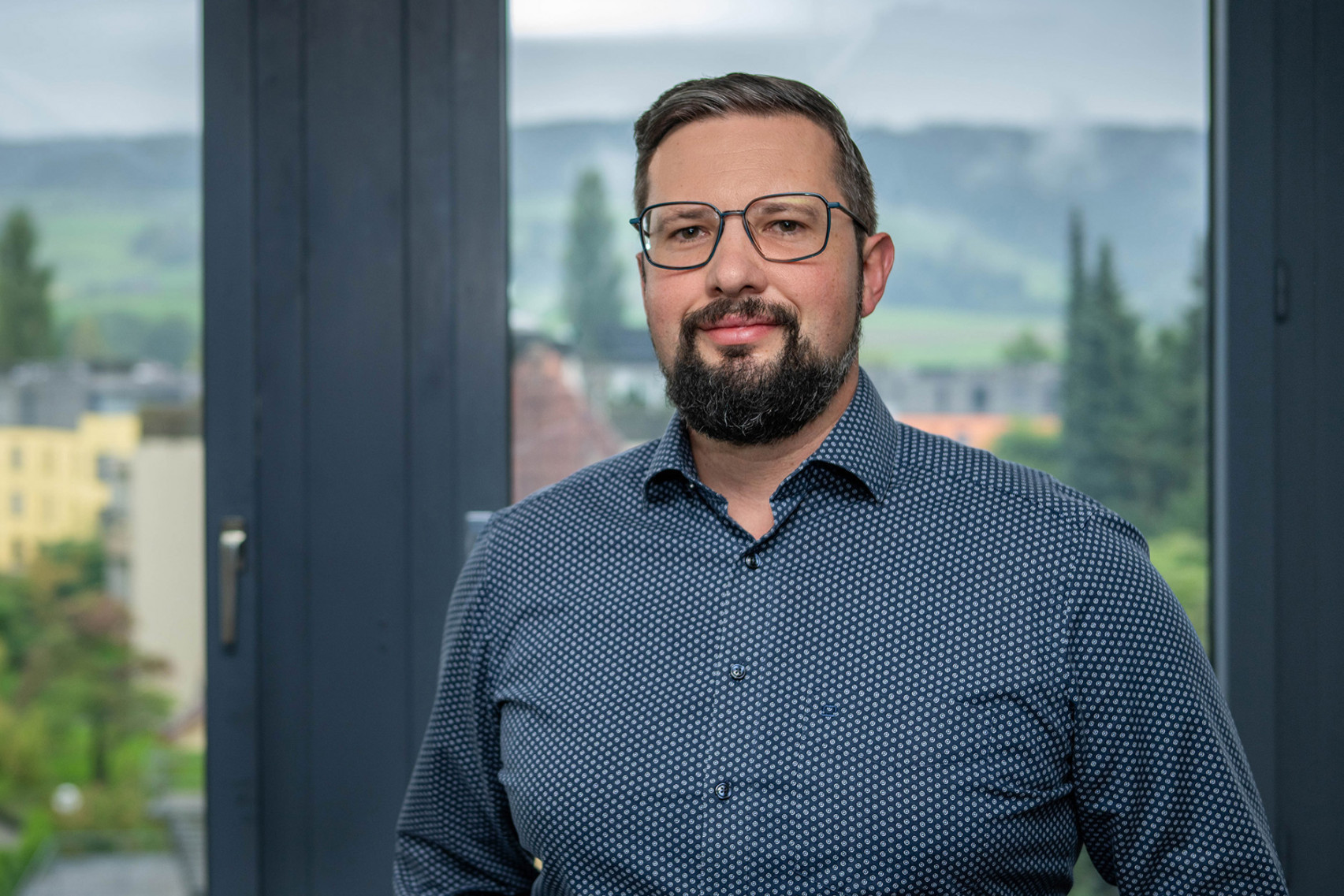 Kundenberater der emmental versicherung steht vor einem Fenster und blickt freundlich in die Kamera.