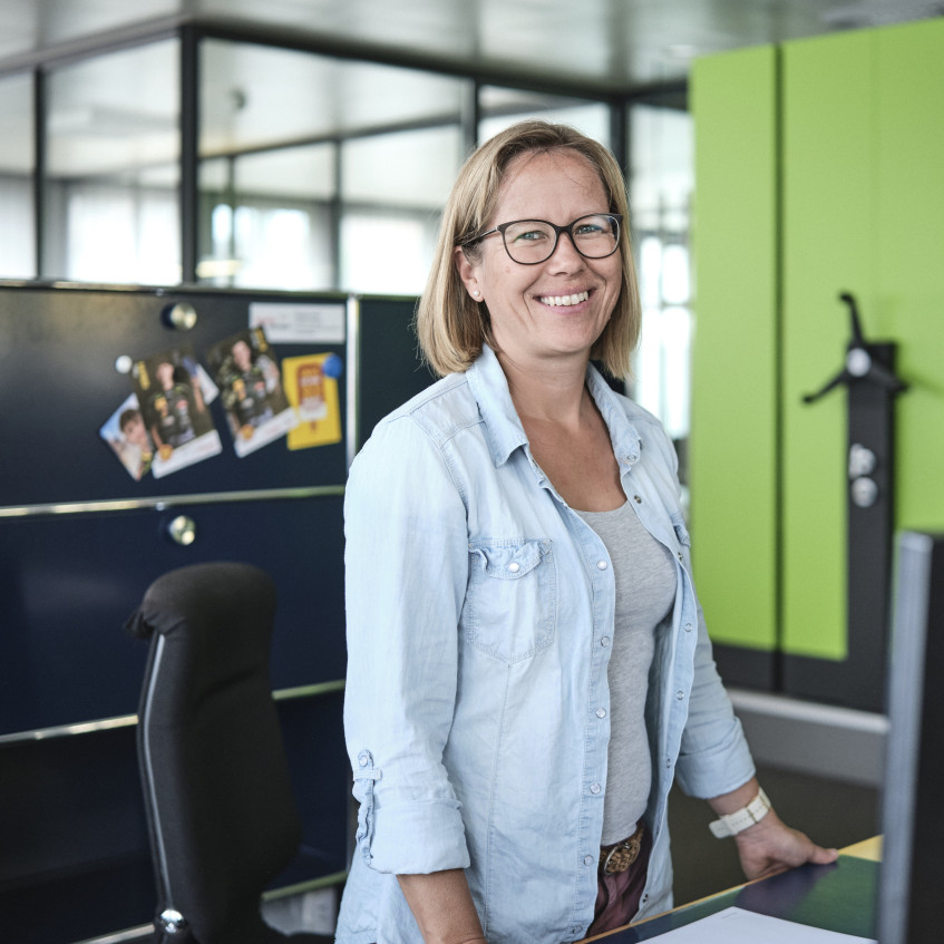 Eine Mitarbeiterin der emmental versicherung steht vor ihrem Arbeitsplatz und lächelt in die Kamera.