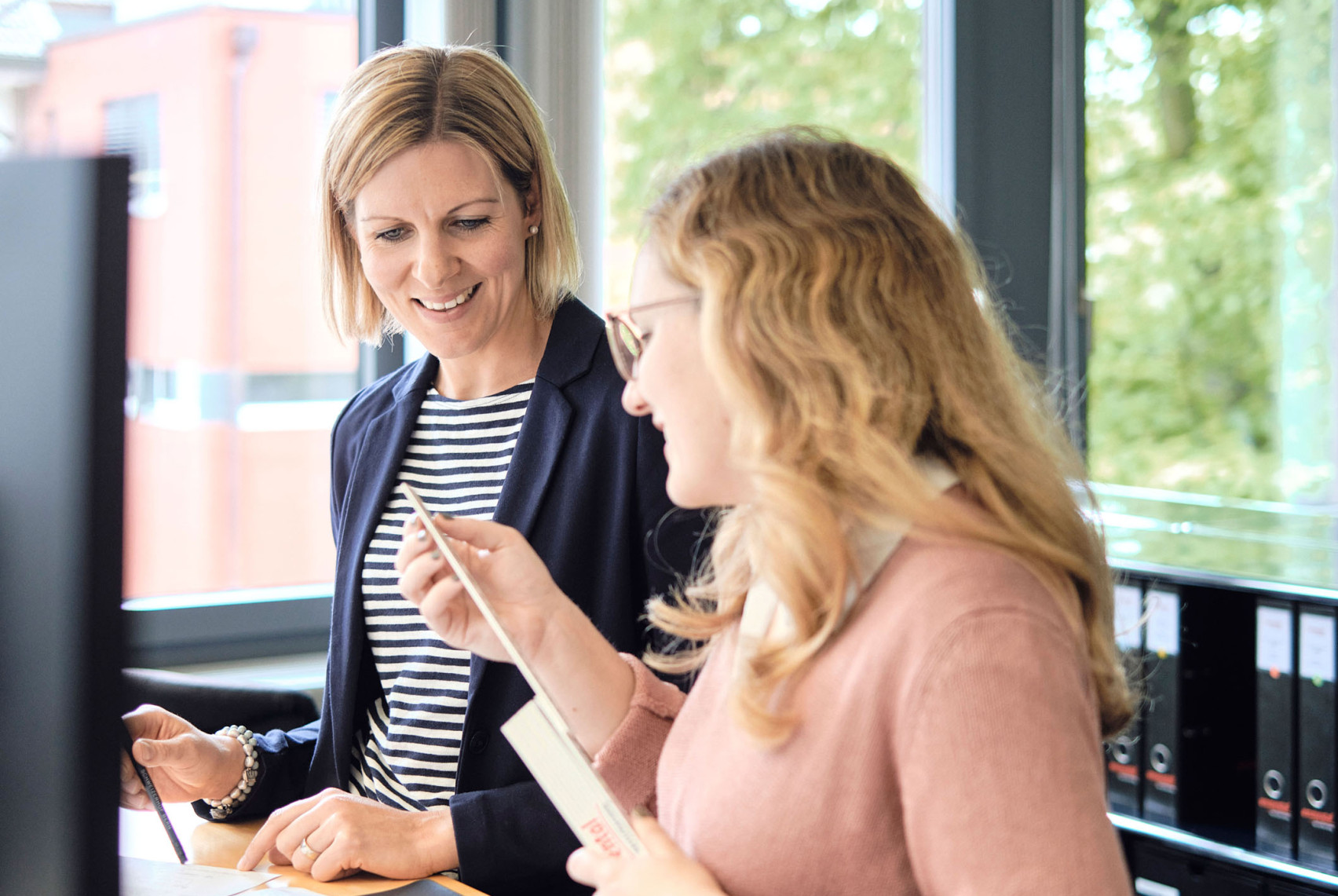 Zwei Mitarbeiterinnen der emmental versicherung stehen vor einem Stehpult, besprechen etwas und lächeln dabei.