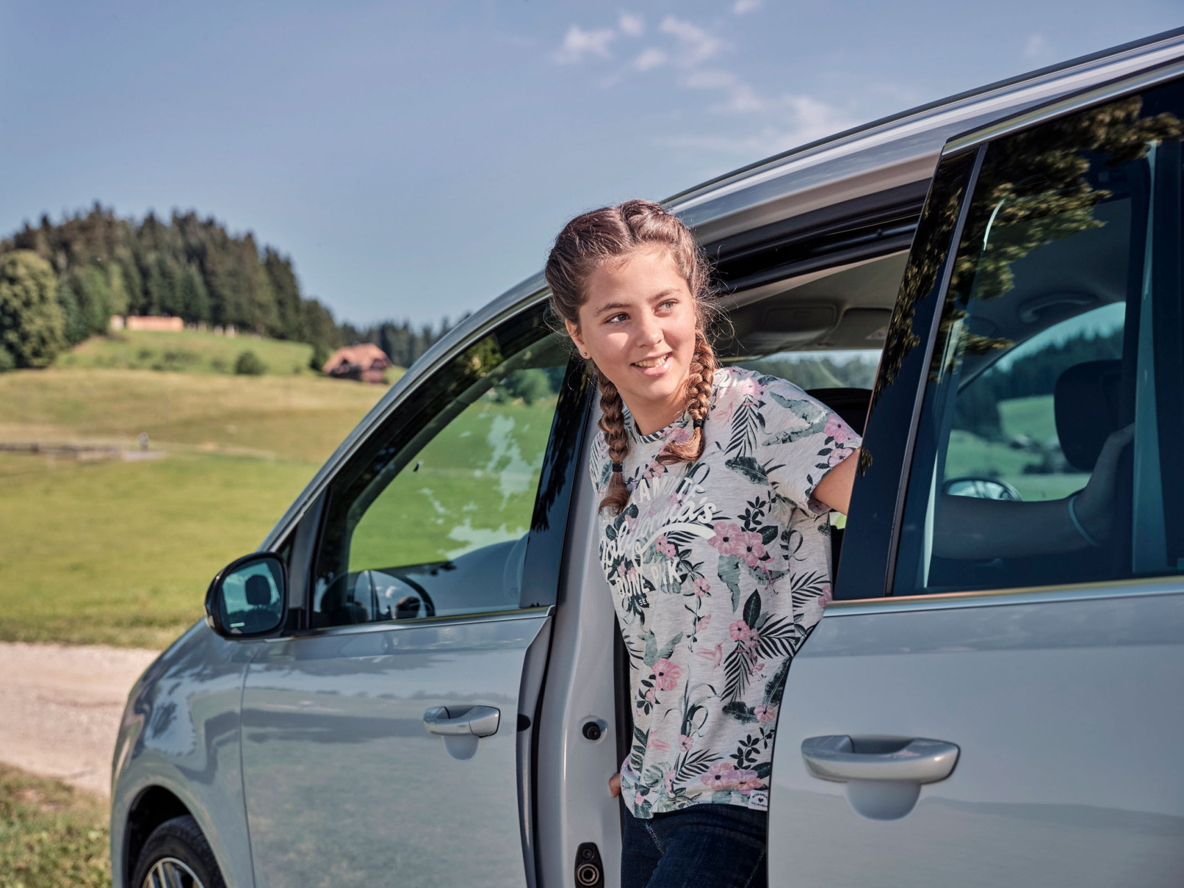 Ein Mädchen im Teenager-Alter steht in der offenen Schiebetür des silbernen Familien-Autos und schaut hinaus. Im Hintergrund sind grüne Felder und Hügel zu sehen.