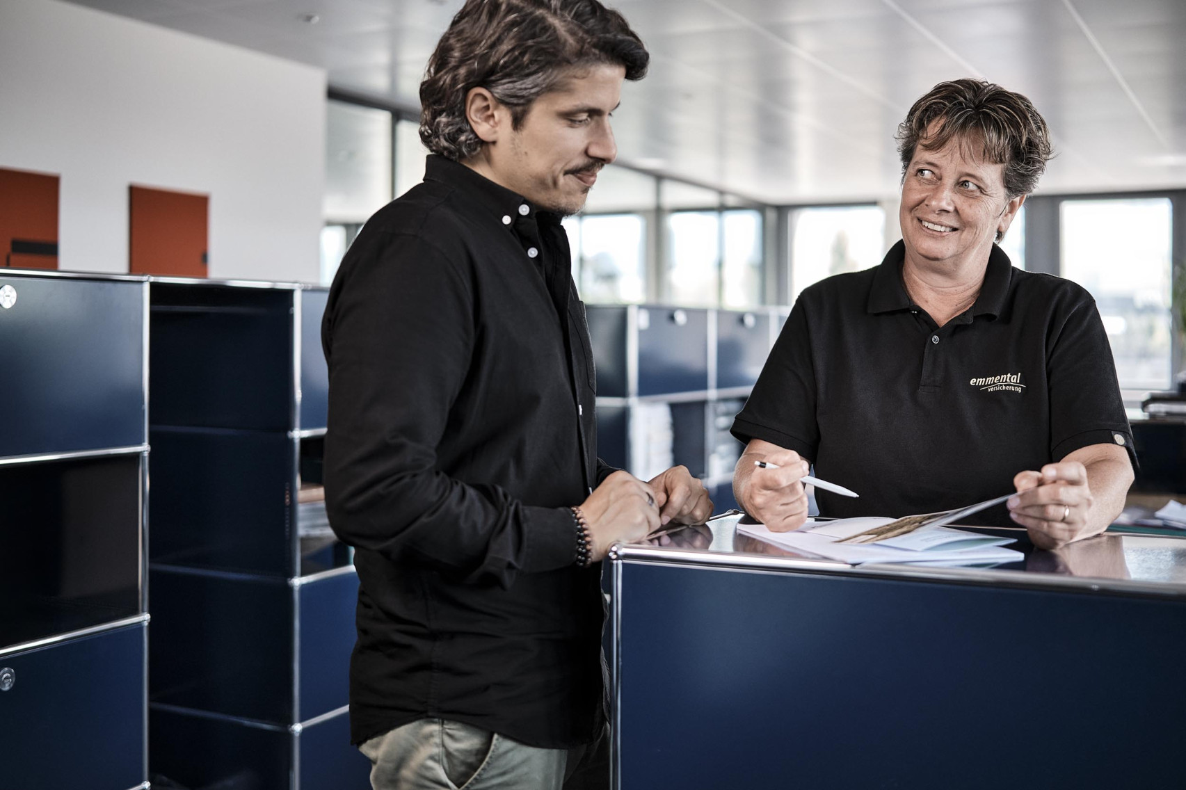 Eine Kundenberaterin der emmental versicherung zeigt einem Kunden etwas in ihrer Produktbroschüre.