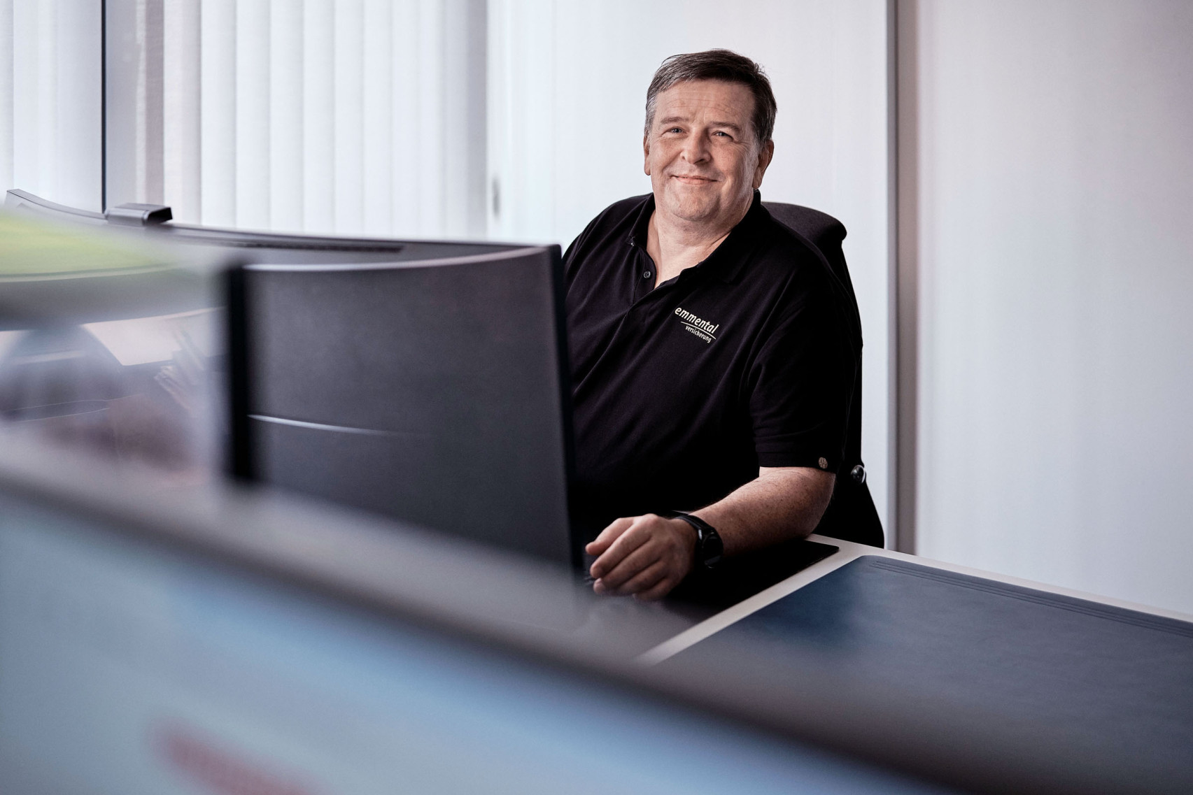 Ein Mitarbeiter der emmental versicherung sitzt an seinem Arbeitsplatz und blickt nebem seinem Bildschirm vorbei lächelnd in die Kamera.