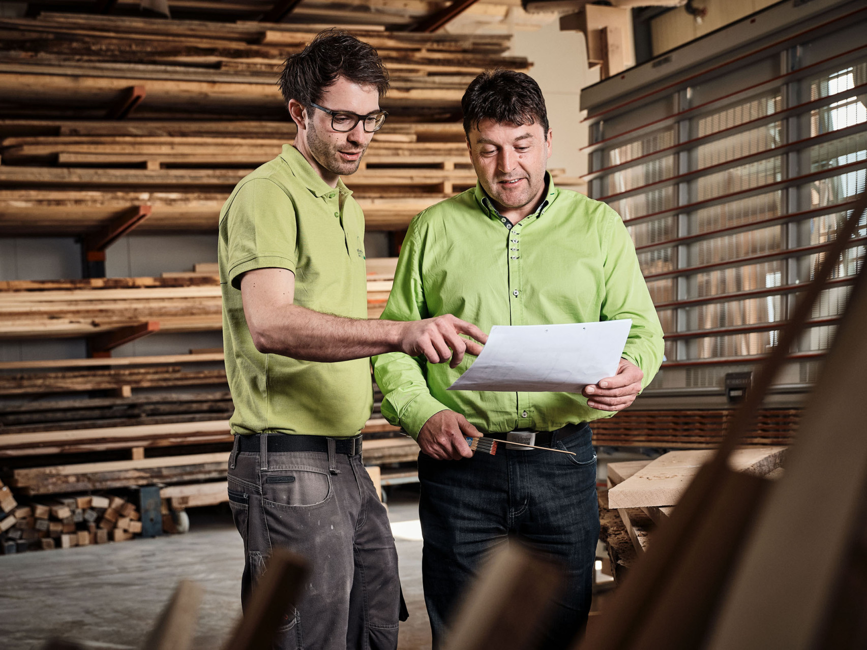 Ein Mitarbeiter einer Schreiner steht mit seinem Vorgesetzten in der Lagerhalle und zeigt ihm ein Dokument.